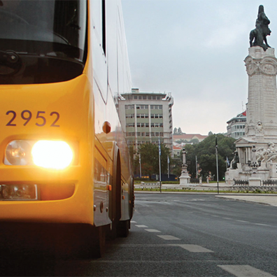Fotografia da frente do autocarro, com detalhe do farol direito aceso, com a estátua do Marquês de Pombal à direita e prédios ao fundo